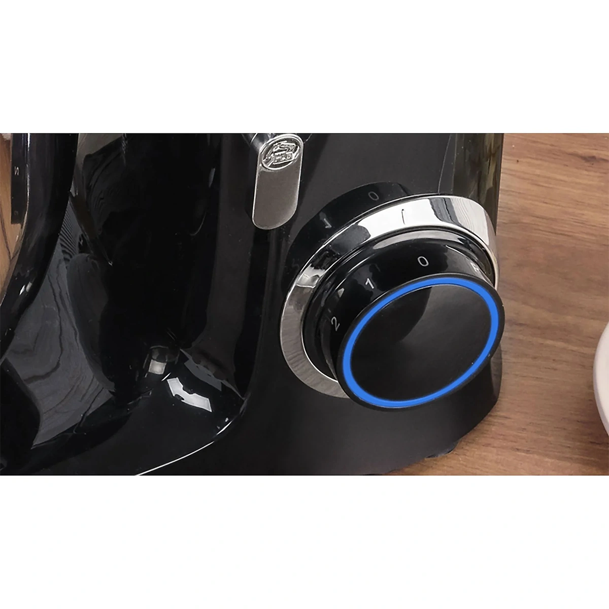 Eine Nahaufnahme der MPM MRK-19 Planeten-Küchenmaschine von MPM mit schwarzem Finish, silbernem und schwarzem, auf Null gestelltem Drehknopf mit blauem LED-Ring, weißer Nummerierung und einem leistungsstarken 2200-Watt-Motor auf einer Holzoberfläche.