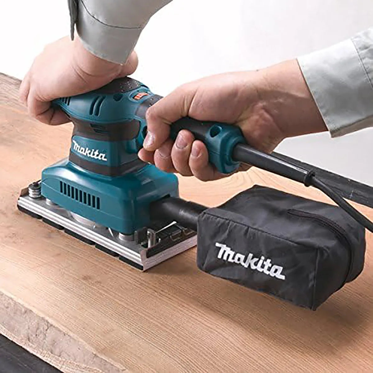 Eine Person glättet Holz mit dem Makita BO3711J Schwingschleifer, der mit einem Staubbeutel ausgestattet ist, das Makita-Logo trägt und in einem MAKPAC Systemkoffer geliefert wird.