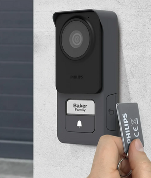 Eine Person hält einen Philips-Schlüsselanhänger in die Nähe einer grauen, an der Wand montierten Video-Türsprechanlage mit der Aufschrift „Baker Family“, bei der es sich um die elegante Video-Türsprechanlage „WelcomeEye Lite 2“ von Philips in Schwarz mit integriertem RFID-Lesegerät handelt.