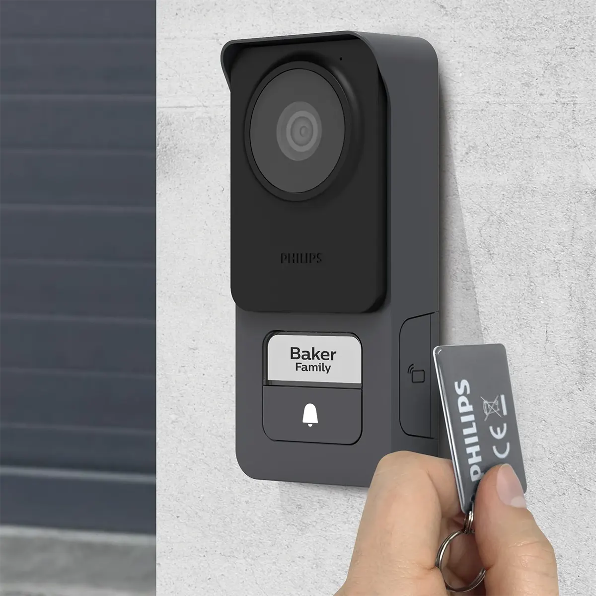 Eine Person hält einen Philips-Schlüsselanhänger in die Nähe einer grauen, an der Wand montierten Video-Türsprechanlage mit der Aufschrift „Baker Family“, bei der es sich um die elegante Video-Türsprechanlage „WelcomeEye Lite 2“ von Philips in Schwarz mit integriertem RFID-Lesegerät handelt.