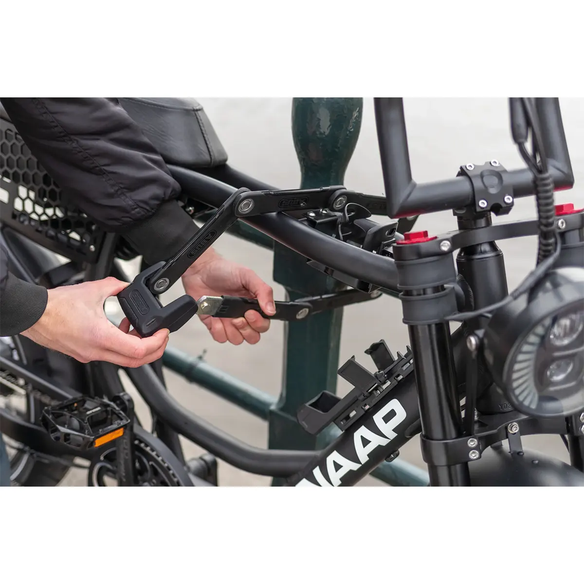 Eine Person sichert ein schwarzes WAAP E-Bike mit dem ABUS Faltschloss Bordo Granit 6500K an einem Pfosten und hält dabei den Schlüssel in der Hand.