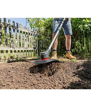 Eine Person bearbeitet die Gartenerde an einem Holzzaun und Grünpflanzen mit der AL-KO TLD 1820 Akku-Bodenhacke - schwarz und genießt die kabellose Bodenbearbeitung mit dem starken AL-KO 18V Akku an einem sonnigen Tag.