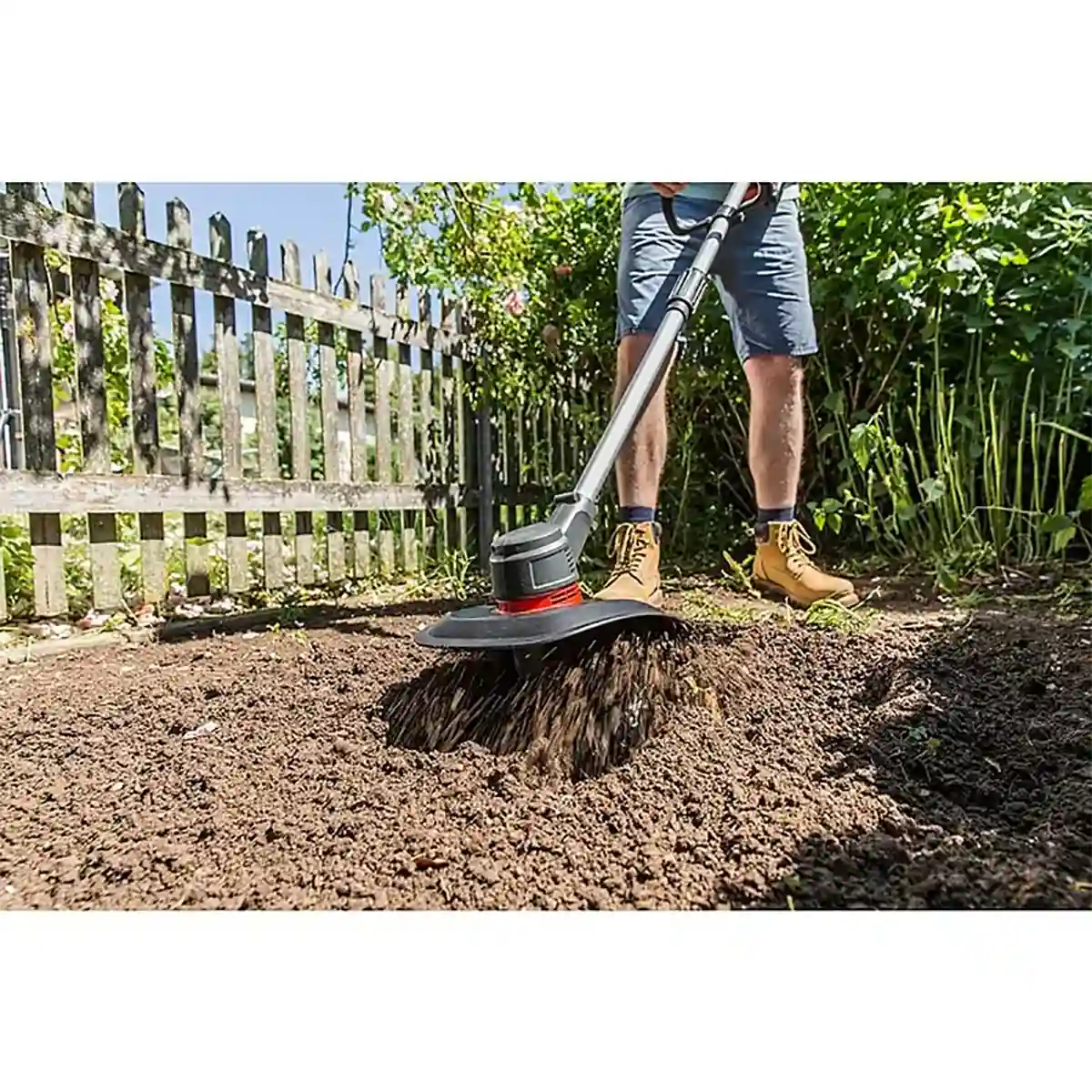 Eine Person bearbeitet die Gartenerde an einem Holzzaun und Grünpflanzen mit der AL-KO TLD 1820 Akku-Bodenhacke - schwarz und genießt die kabellose Bodenbearbeitung mit dem starken AL-KO 18V Akku an einem sonnigen Tag.