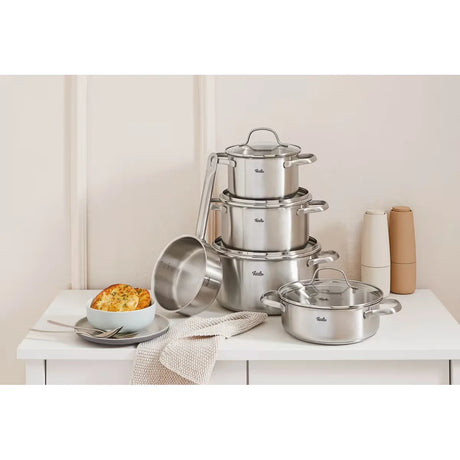 Das Fissler Kochtopfset TORONTO 5-teilig aus Edelstahl steht mit Deckeln auf einem weißen Tisch, neben einer Schüssel mit Essen, Serviette sowie Salz- und Pfefferstreuern; für Induktion geeignet.