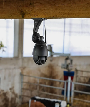 Die Kerbl IPCam 10809 Sicherheitskamera - schwarz, eine WLAN-Kamera mit Nachtsicht, ist an einem Holzbalken in einer Scheune installiert, in der Vieh und landwirtschaftliche Geräte sichtbar sind.