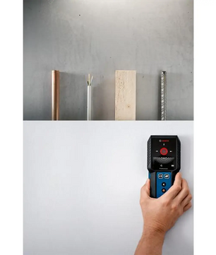 Eine Hand bedient das Ortungsgerät GMS 120-27 (blau) von Bosch Professional auf einer weißen Oberfläche, wobei oben die erkannten Objekte wie Rohre (Leitungen erkennen), Holz und Bewehrungsstahl angezeigt werden.
