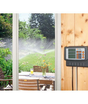 An einer Holzwand neben der Terrasse ist eine GARDENA Classic 4030 Bewässerungssteuerung – grau angebracht, die die Gartenbewässerung automatisch steuert, während Sprinkler den Rasen draußen bewässern.
