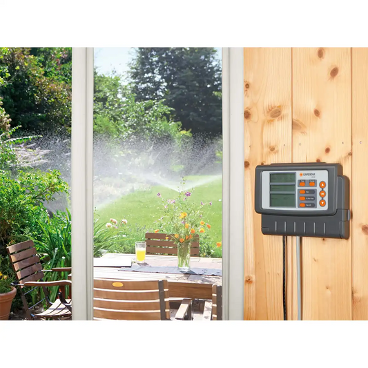 An einer Holzwand neben der Terrasse ist eine GARDENA Classic 4030 Bewässerungssteuerung – grau angebracht, die die Gartenbewässerung automatisch steuert, während Sprinkler den Rasen draußen bewässern.
