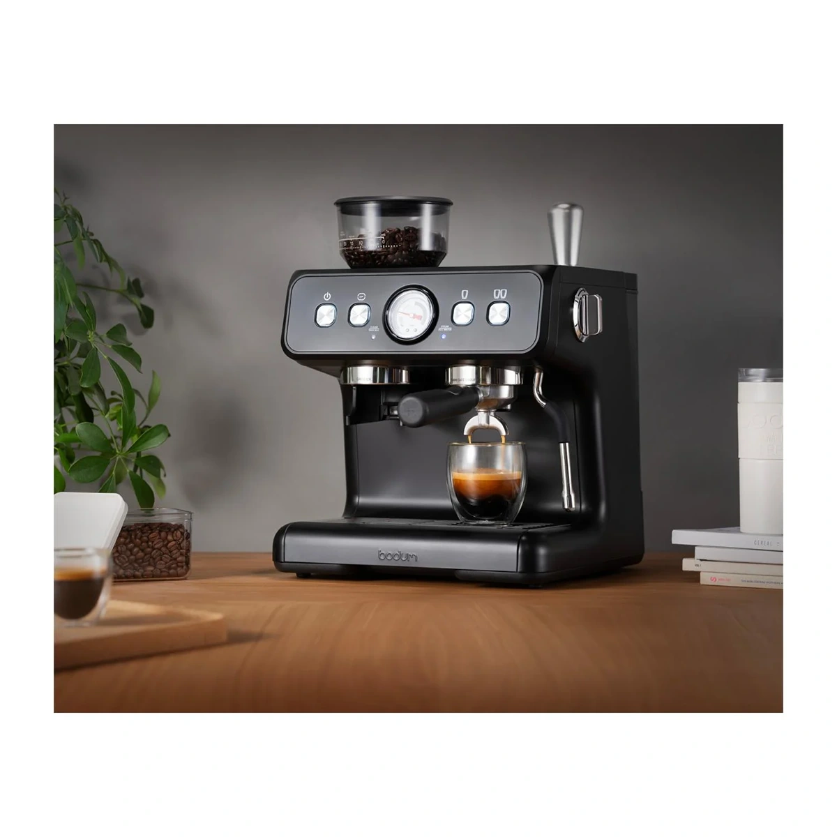 Die Bodum Bistro 12459-01 Espressomaschine in Schwarz von Bodum bereitet aromatischen Espresso auf einer Holztheke zu - umgeben von Kaffeebohnen, Tassen und einer Pflanze, ideal für frisch gebrühten Espresso und feinen Milchschaum.