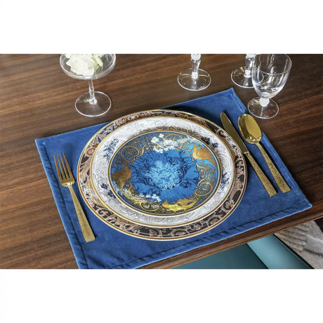 Elegantes blau-goldenes Tafelservice auf blauem Tischset, ergänzt durch Sambonet Siena PVD Gold Besteckset (24-teilig) und Klarglas auf einem Holztisch.