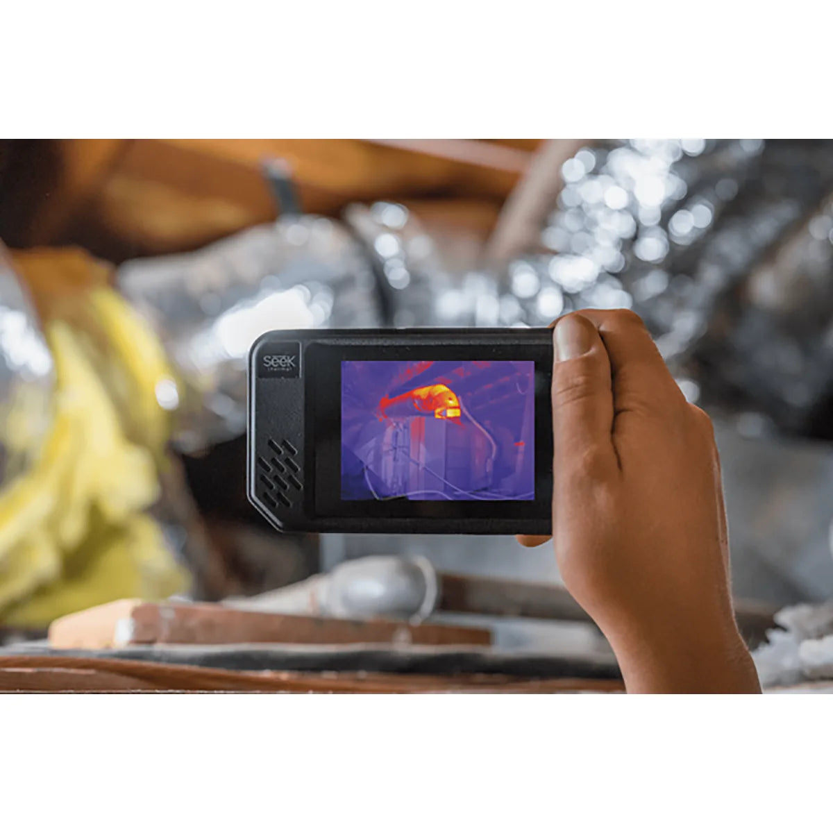 Eine Hand hält eine Seek Thermal SHOT PRO Wärmebildkamera von Seek Thermal in Schwarz, die die Wärmesignaturen von Objekten in einem Dachgeschoss aufnimmt.