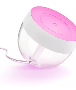 Die Philips Hue Ambiance Iris Tischleuchte – weiß von Philips besticht durch ihr rundes Design mit einer rosa Oberseite und strahlt ein sanftes rosa Licht aus, das für eine gemütliche Stimmungsbeleuchtung sorgt, wenn sie über ein weißes Kabel an die Steckdose angeschlossen ist.