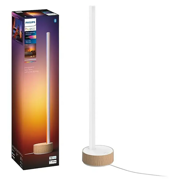 Die hohe, schlanke Tischleuchte „Philips Hue Gradient Signe“ – weiß von Philips mit Holzsockel steht neben ihrer Verpackung in Sonnenuntergangsfarben und verfügt über die innovative Gradient-Technologie für eine intelligente Lichtsteuerung.