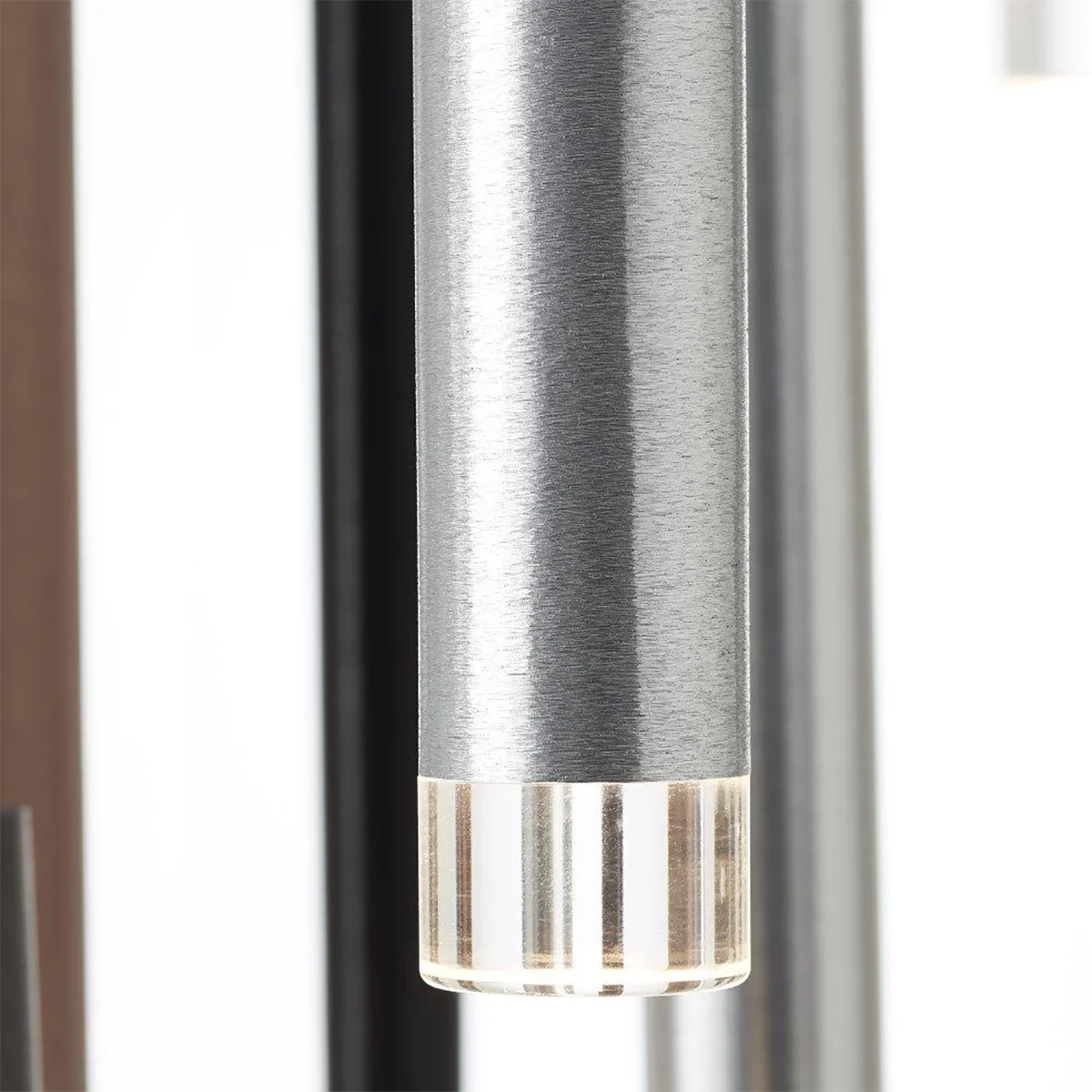 Eine Nahaufnahme der Brilliant Living Cembalo 16-flammig LED-Pendelleuchte in Braun/Kaffee, die sich durch Energieeffizienz, dimmbare Beleuchtung und eine gebürstete silberne Oberfläche mit einem transparenten, geriffelten Ende vor einem verschwommenen vertikalen Hintergrund auszeichnet.