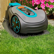 Der GARDENA 15202-20 SILENO minimo Mähroboter dockt auf dem Rasen an einem Holzzaun und rosa Blumen an und bietet innovative Rasenpflege mit smarter Bluetooth-App-Steuerung.