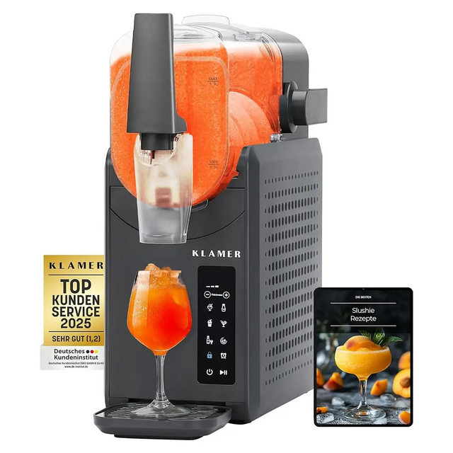 Die KLAMER Slush-Maschine mit 2,6 l – grau von KLAMER, die orangefarbenen Slush in ein Glas füllt, daneben liegen ein Rezeptbuch und eine Auszeichnungsplakette.
