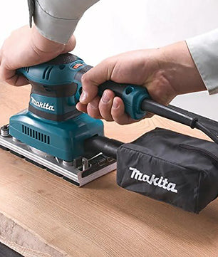 Eine Person glättet Holz mit dem Makita BO3711J Schwingschleifer, der mit einem Staubbeutel ausgestattet ist, das Makita-Logo trägt und in einem MAKPAC Systemkoffer geliefert wird.