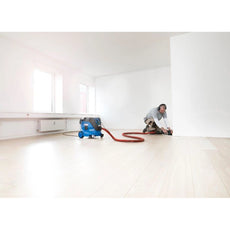 Eine Person mit Schutzkopfhörern schleift einen Boden an einer weißen Wand in einem hellen, leeren Raum. Ein Nilfisk ATTIX 33-2M PC Sicherheitssauger - M-Klasse, 30 l, 1400 W ist zur Staubabsaugung an den Schleifer angeschlossen.