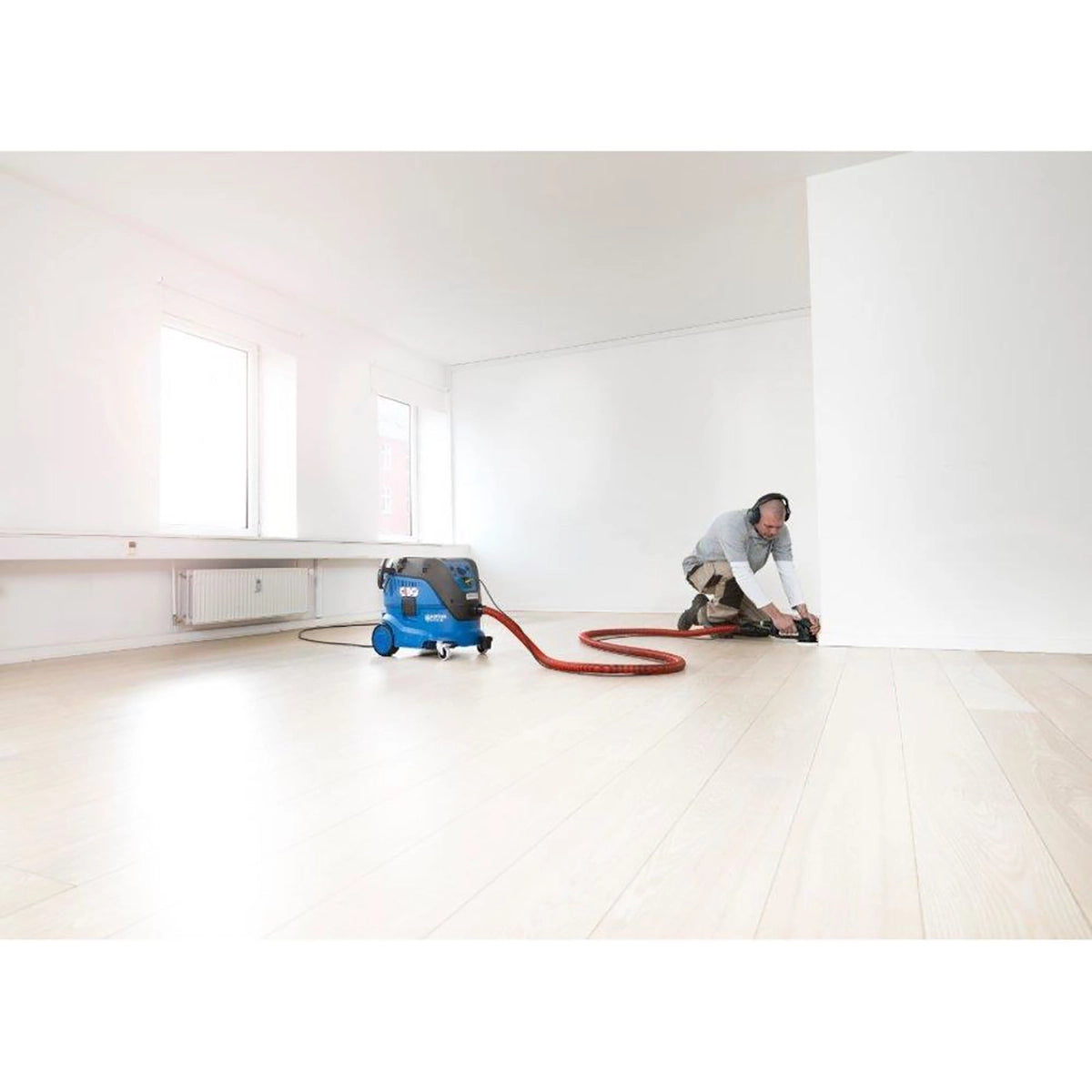 Eine Person mit Schutzkopfhörern schleift einen Boden an einer weißen Wand in einem hellen, leeren Raum. Ein Nilfisk ATTIX 33-2M PC Sicherheitssauger - M-Klasse, 30 l, 1400 W ist zur Staubabsaugung an den Schleifer angeschlossen.