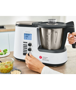 Eine Person nimmt die Einstellungen an einer SILVERCREST Monsieur Cuisine edition plus SKMK 1100 A1 Küchenmaschine vor, die auf einer Arbeitsplatte steht, umgeben von Gemüse, einer Schüssel mit Lebensmitteln und frischen Zutaten.