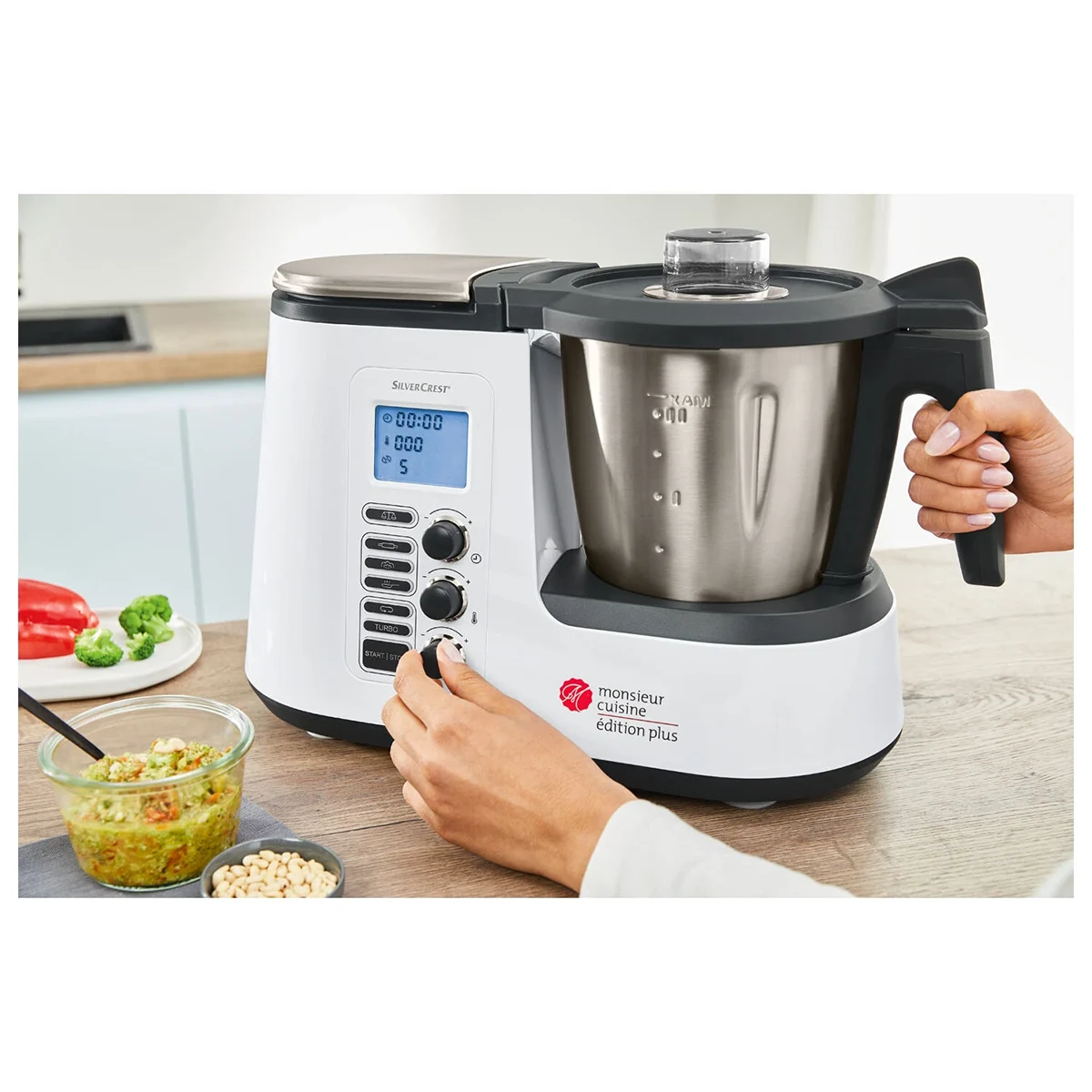 Eine Person nimmt die Einstellungen an einer SILVERCREST Monsieur Cuisine edition plus SKMK 1100 A1 Küchenmaschine vor, die auf einer Arbeitsplatte steht, umgeben von Gemüse, einer Schüssel mit Lebensmitteln und frischen Zutaten.