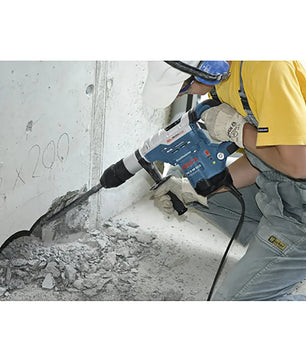 Ein Arbeiter in Schutzkleidung verwendet den Bosch GBH 5-40 DCE Professional Bohrhammer mit SDS-max von Bosch, um eine Betonwand zu durchbrechen, wobei Trümmer auf dem Boden sichtbar sind.