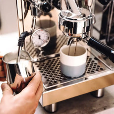 Nahaufnahme der Hände beim Aufbrühen von Milch und Espresso mit der Bezzera BZ10 PM Zweikreiser Espressomaschine aus Edelstahl, mit einer präzisen Brühgruppe, von Bezzera, neben einer Tasse.