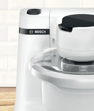 Nahaufnahme einer weißen Bosch BOSCH Küchenmaschine - MUMS2AW01 auf einer Holztheke, mit klarer Rührschüssel, schwarzen Akzenten und fortschrittlichem 3D PlanetaryMixing für perfekte Ergebnisse.