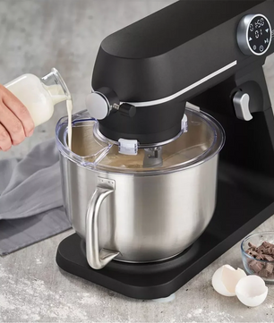 Eine Hand gießt Flüssigkeit aus einem kleinen Krug in die schwarze CASO KM 1800 Küchenmaschine mit einer 6-Liter-Edelstahlschüssel voller Teig. Eierschalen, Schokoladenstücke und ein graues Tuch liegen auf dem hellen Tresen daneben.