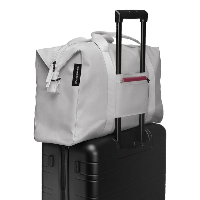 Ein Horizn Studios SoFo Weekender M - Light Quartz Grey aus recycelter Baumwolle hängt an einem schwarzen Rollkoffer, der Griffe über den ausgezogenen Griff legt - eine stilvolle, nachhaltige Reisetasche von Horizn Studios.