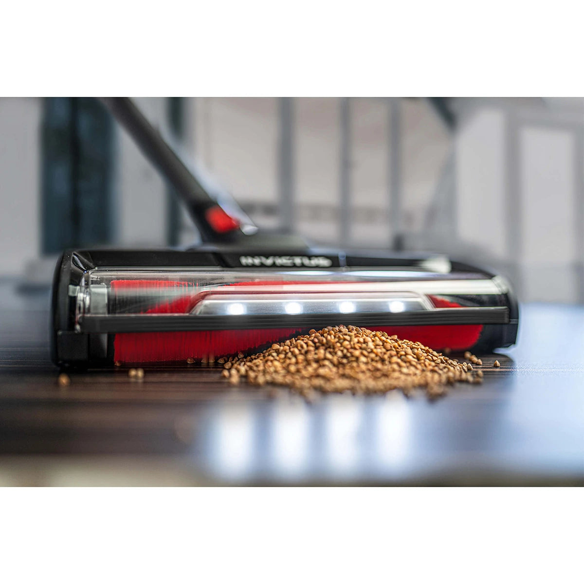 Eine Nahaufnahme des INVICTUS X9 Akkustaubsaugers in schwarz von INVICTUS mit LED-Beleuchtung und HEPA-Filter bei der Reinigung von verschütteten Körnern auf einem dunklen Hartholzboden in Innenräumen.