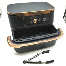 Eine schwarz-goldene Ninja Foodi FlexDrawer Dual Zone Heißluftfritteuse von Ninja mit zwei 4-Liter-Körben wird mit einer Metall-Küchenzange davor gezeigt.