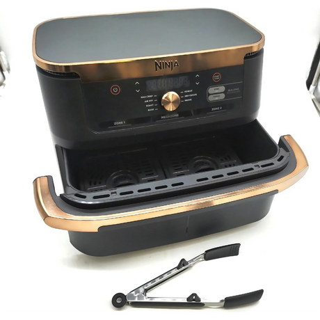 Eine schwarz-goldene Ninja Foodi FlexDrawer Dual Zone Heißluftfritteuse von Ninja mit zwei 4-Liter-Körben wird mit einer Metall-Küchenzange davor gezeigt.