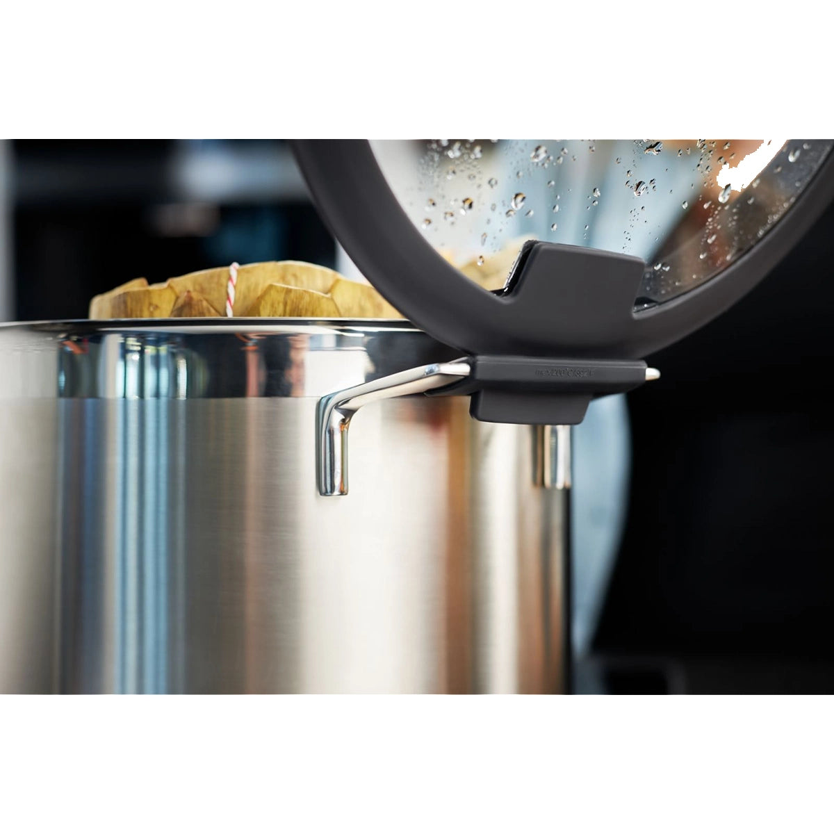 Eine Nahaufnahme eines Rösle Silence Pro Topfset 6-tlg. Edelstahltopfes von Rösle, mit einem auf dem Henkel ruhenden Glasdeckel, Wassertropfen auf dem Deckel und Holzutensilien in diesen hochwertigen Edelstahl-Töpfen.