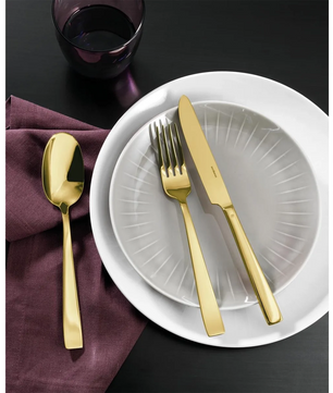 Das Sambonet Flat Besteck 24tlg. - gold von Sambonet, aus Edelstahl 18/10 mit PVD-Gold-Finish, ruht auf einer violetten Serviette neben einem weißen Teller mit durchscheinendem grauen Innenteller und goldenem Besteck. Darüber steht ein dunkles Glas auf dem schwarzen Tisch.