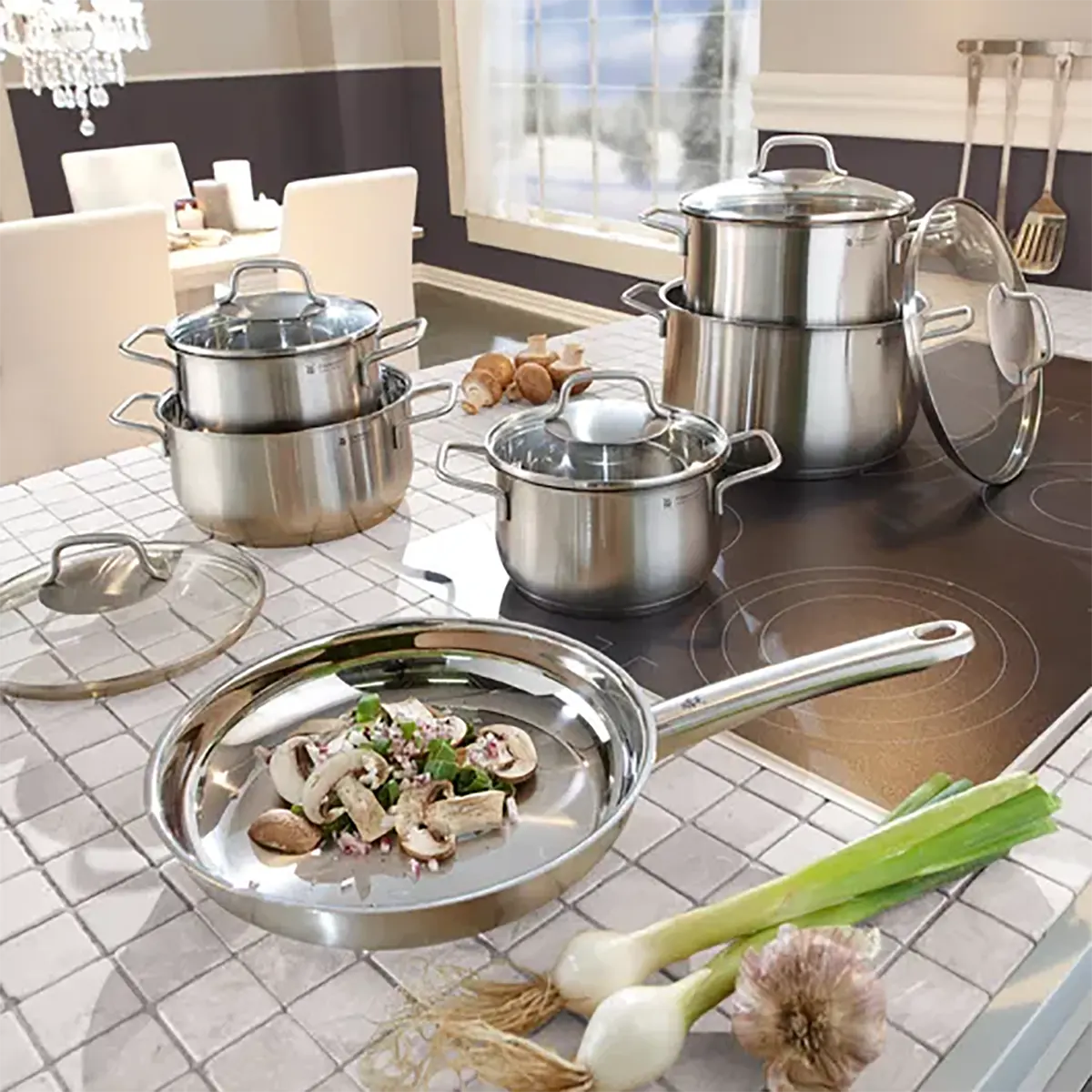 Das 6-teilige WMF Brillant Kochgeschirr-Set aus Edelstahl mit Glasdeckeln steht auf einer modernen Küchenarbeitsplatte neben Frühlingszwiebeln, Knoblauch und einem Schneidebrett mit geschnittenem Gemüse im Vordergrund.